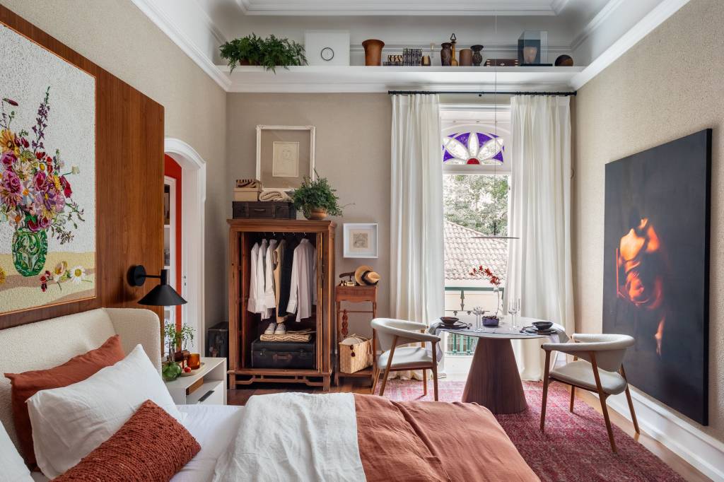 Suíte ganha conforto quarto de hotel com paredes de madeira. Projeto de Paula Muller para a CASACOR Rio 2023. Na foto, espaço de refeições com quadro na parede e mesa redonda. Cama de casal e parede de madeira.