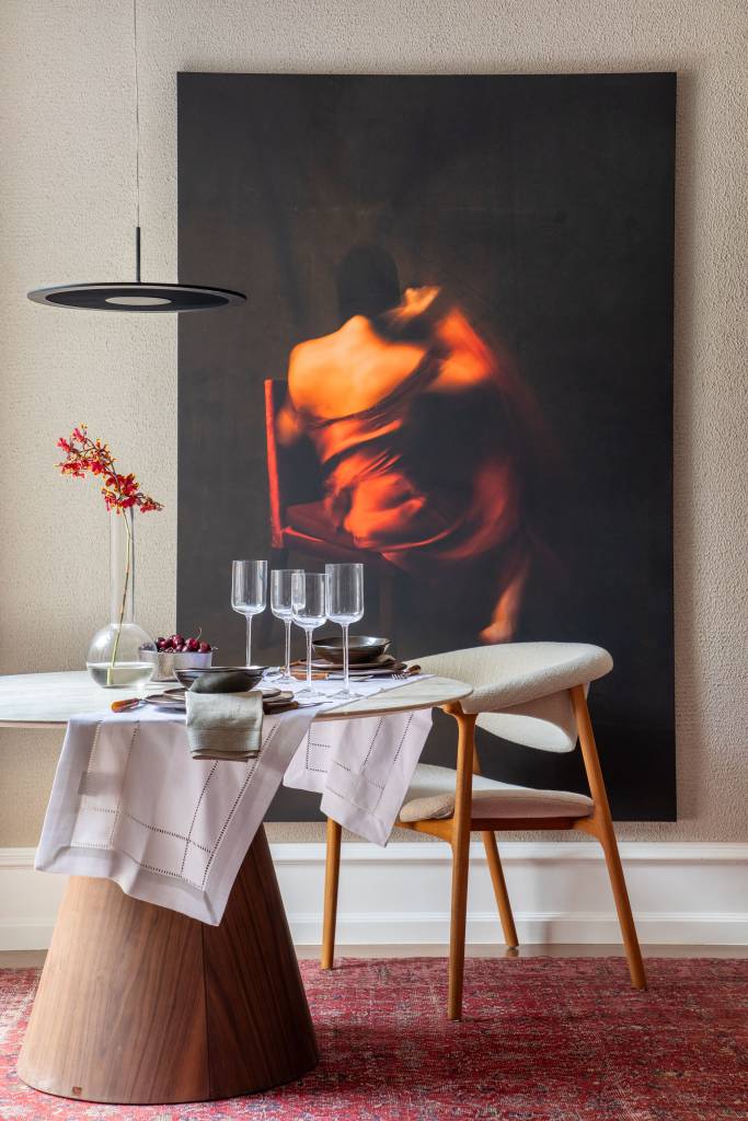 Suíte ganha conforto quarto de hotel com paredes de madeira. Projeto de Paula Muller para a CASACOR Rio 2023. Na foto, espaço de refeições com quadro na parede e mesa redonda.