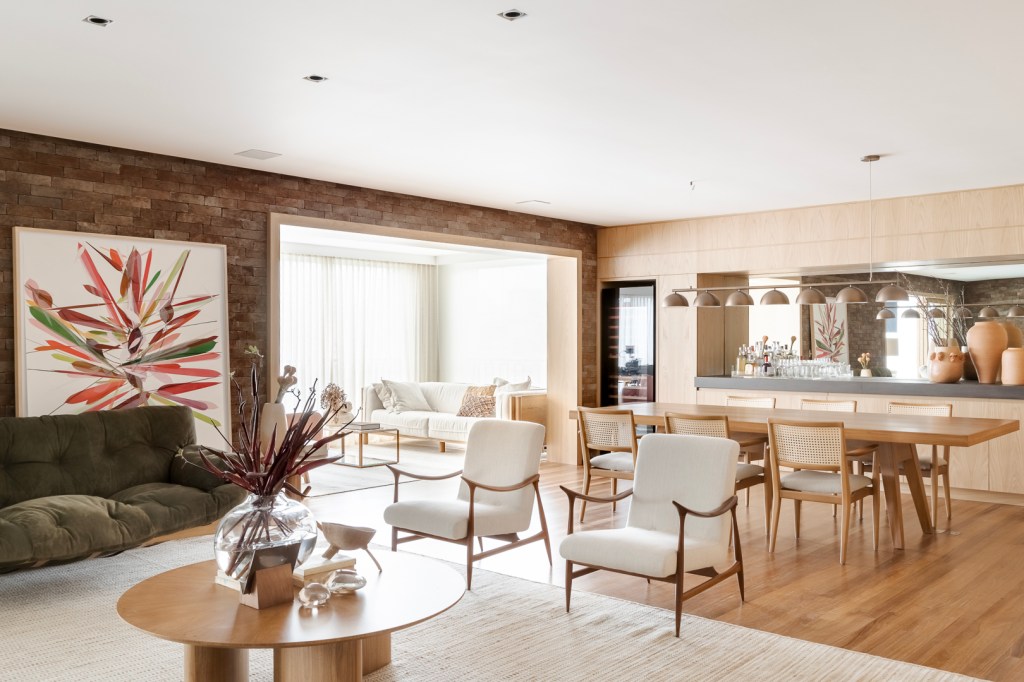 Tijolinhos revestem todas as paredes do living deste apê de 310 m². Projeto de Très Arquitetura. Na foto, sala de estar e jantar com parede de madeira e poltronas.