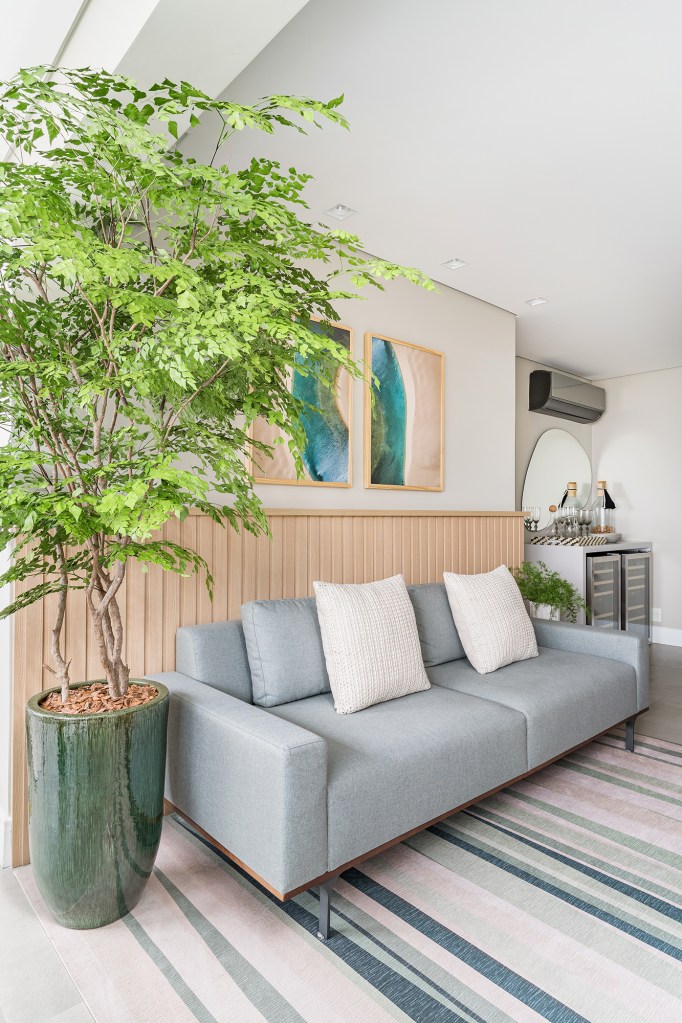 Apartamento de 60 m² ganha décor delicado repleto de madeira e plantas. Projeto de Bia Hajnal. Na foto, sala de estar com sofá cinza e lambri de madeira., Arvore em vaso.