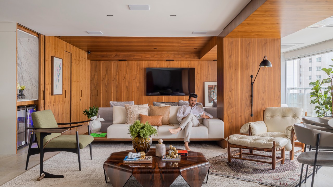Projeto de Romário Rodrigues. Na foto, sala de estar com parede revestida de madeira, poltrona mole e mesa de centro de madeira.