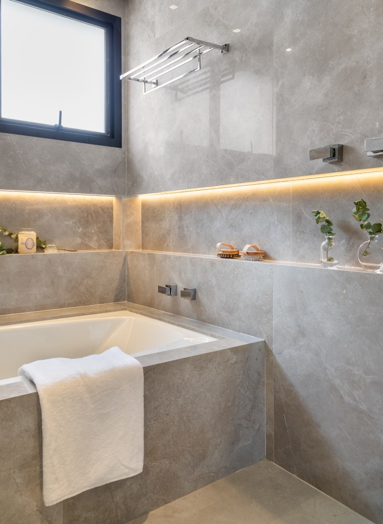 Projeto de Fernanda Dabbur. Na foto, banheiro com revestimentos cinzas, banheira e nicho no box iluminado.