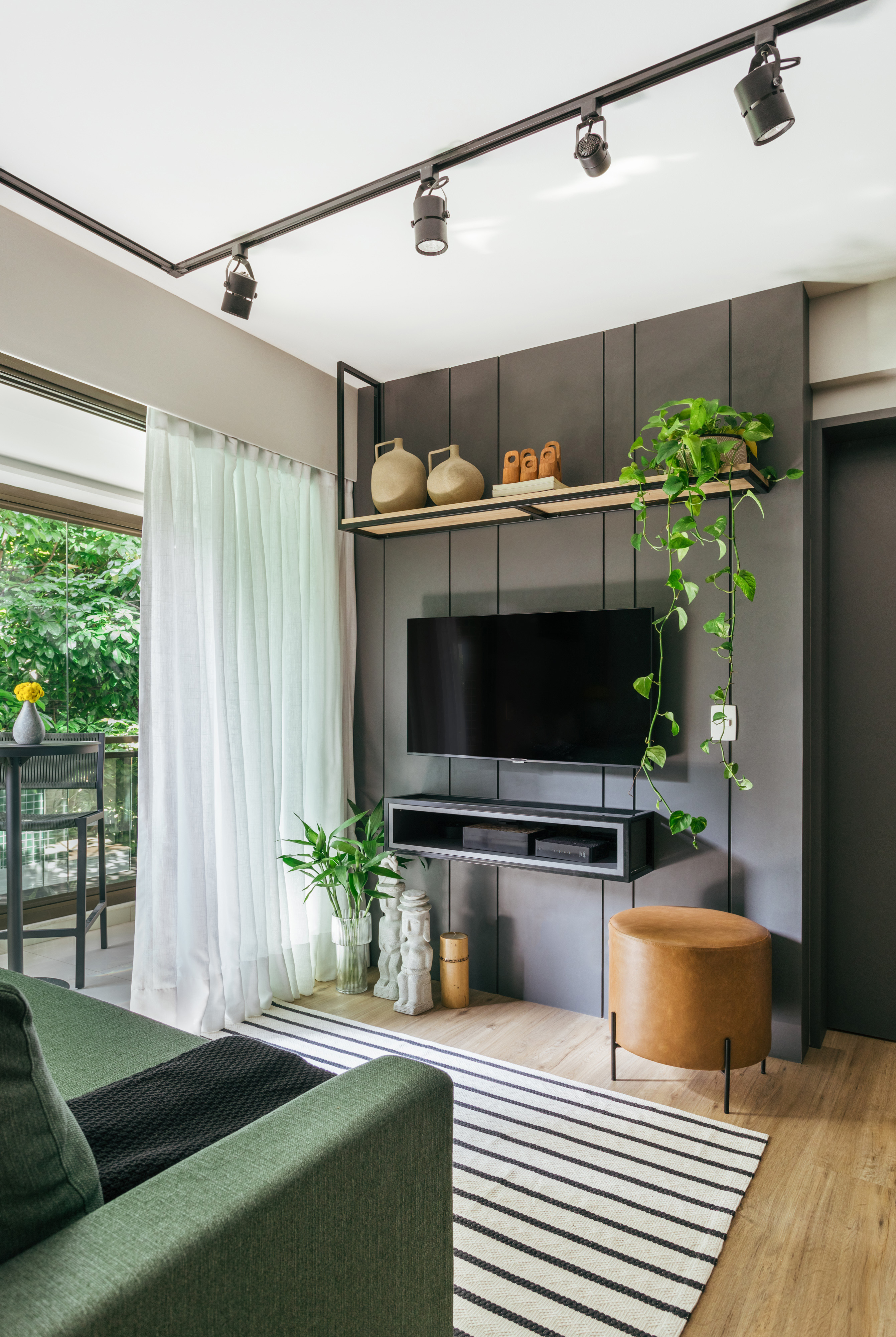 Projeto de Ikeda Arquitetura. Na foto, sala de estar com parede de marcenaria cinza, tv, pufe pequeno e prateleira. Iluminação com trilhos de spots e sofá verde.