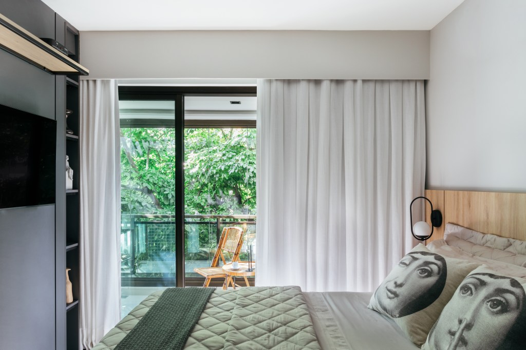 Projeto de Ikeda Arquitetura. Na foto, quarto com cama de casal, cortina branca, painel de madeira cinza escura e porta para varanda pequena.