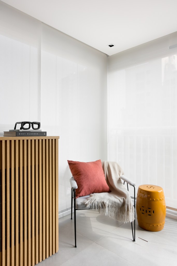Projeto de Studio Dyo. Na foto, varanda integrada com persiana branca e poltrona com manta, almofada e pufe pequeno.