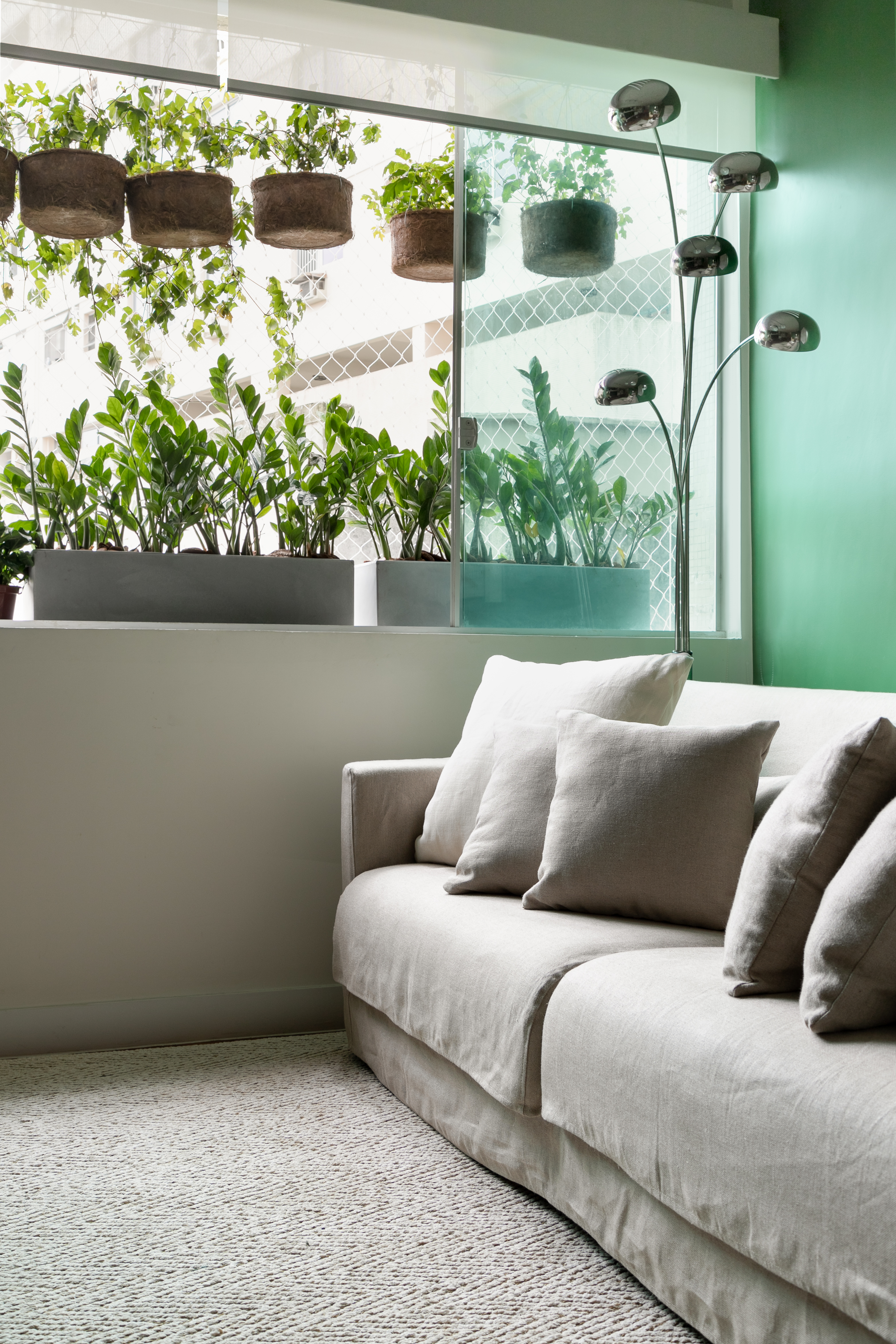 Projeto de Marcela Martins. Na foto, sala de estar com sofá claro, parede verde e plantas na janela.