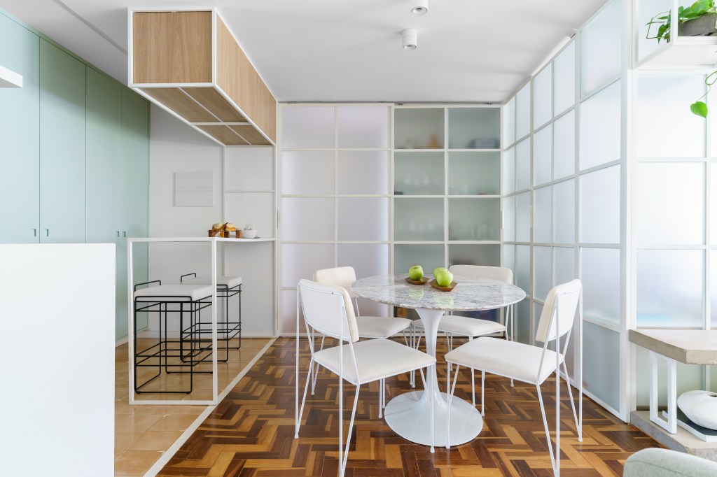 Projeto de Traama Arquitetura. Na foto, sala de jantar integrada com cozinha com piso de taco, mesa e cadeiras brancas e painel de vidro como divisória.