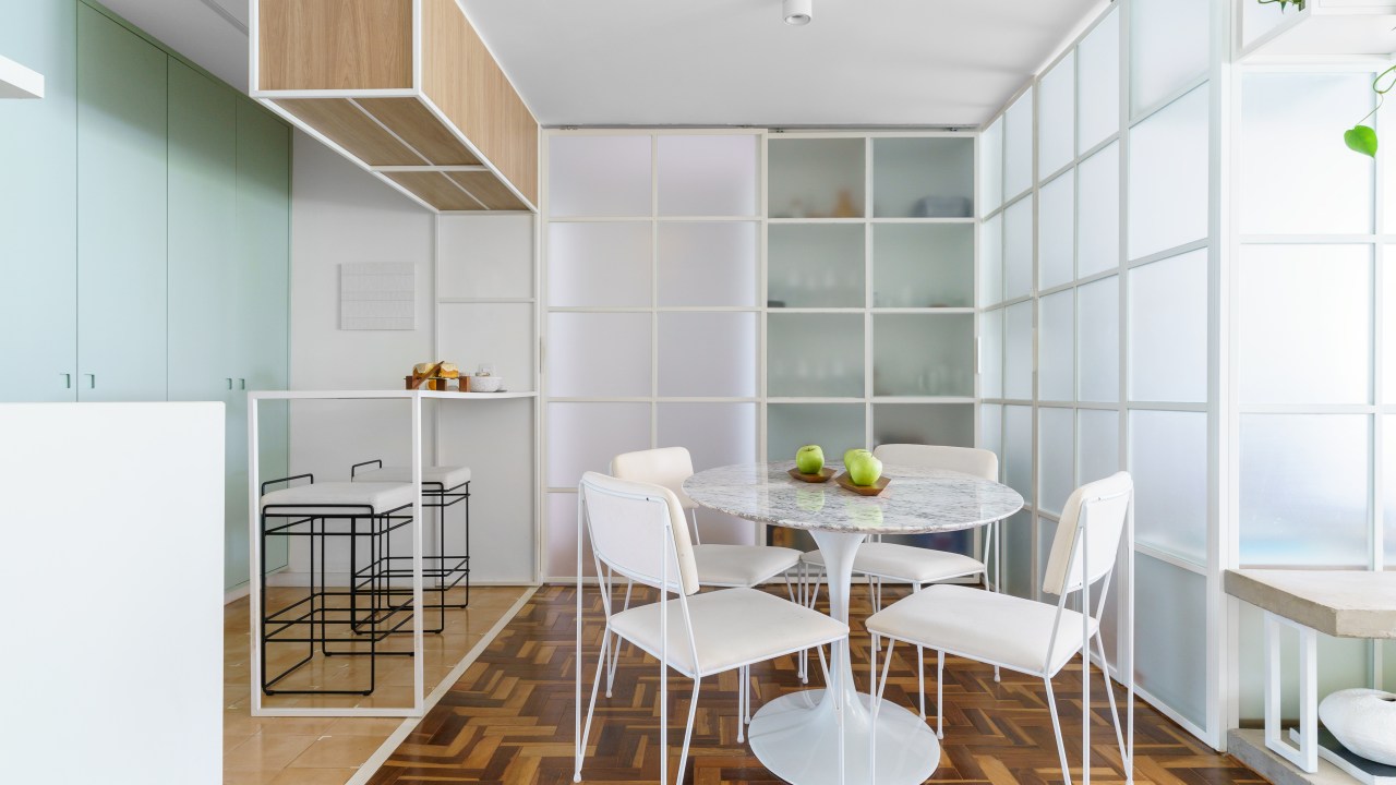 Projeto de Traama Arquitetura. Na foto, sala de jantar integrada com cozinha com piso de taco, mesa e cadeiras brancas e painel de vidro como divisória.