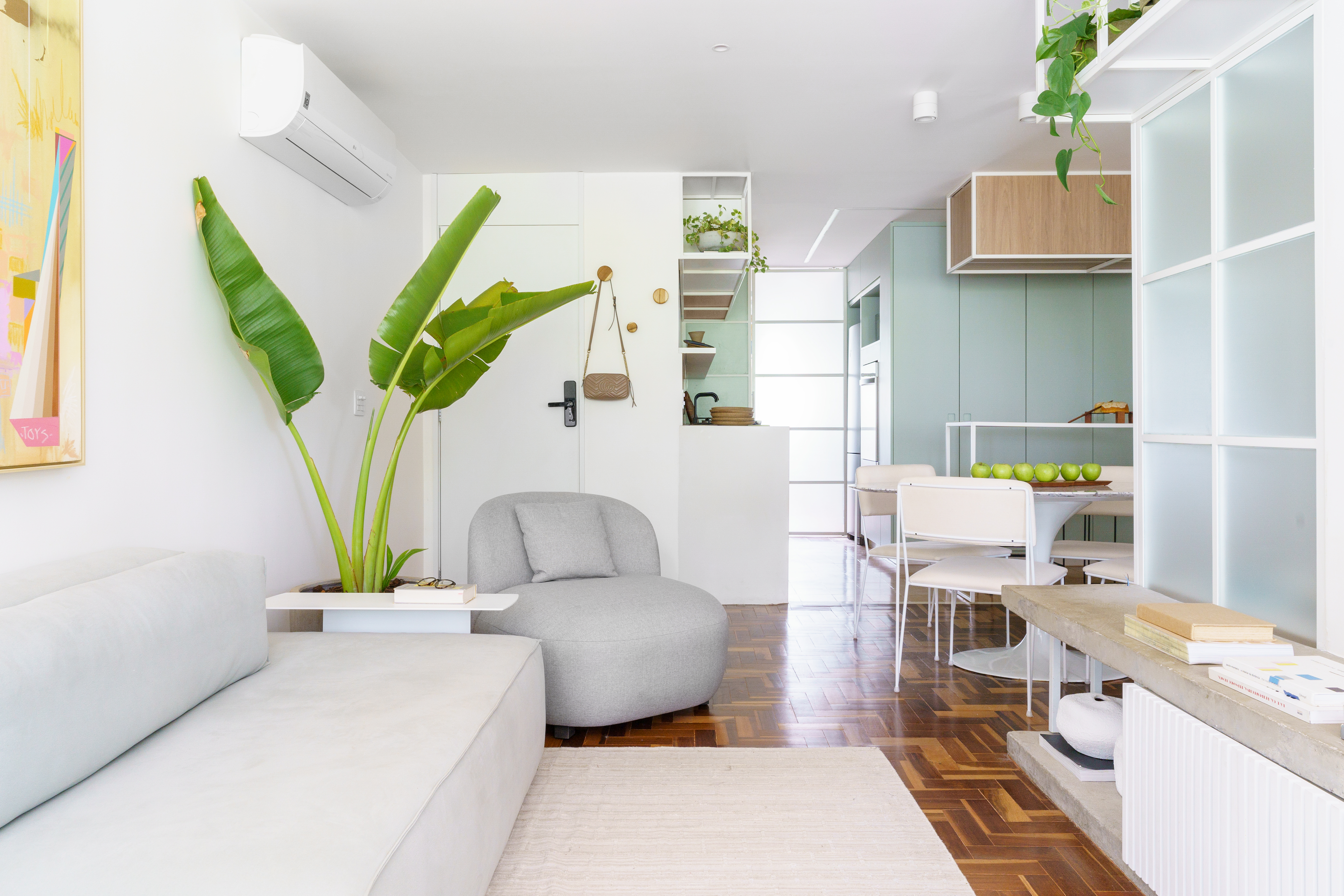 Projeto de Traama Arquitetura. Na foto, sala de estar integrada com piso de taco, poltrona cinza, sofá cinza claro e planta bananeira.