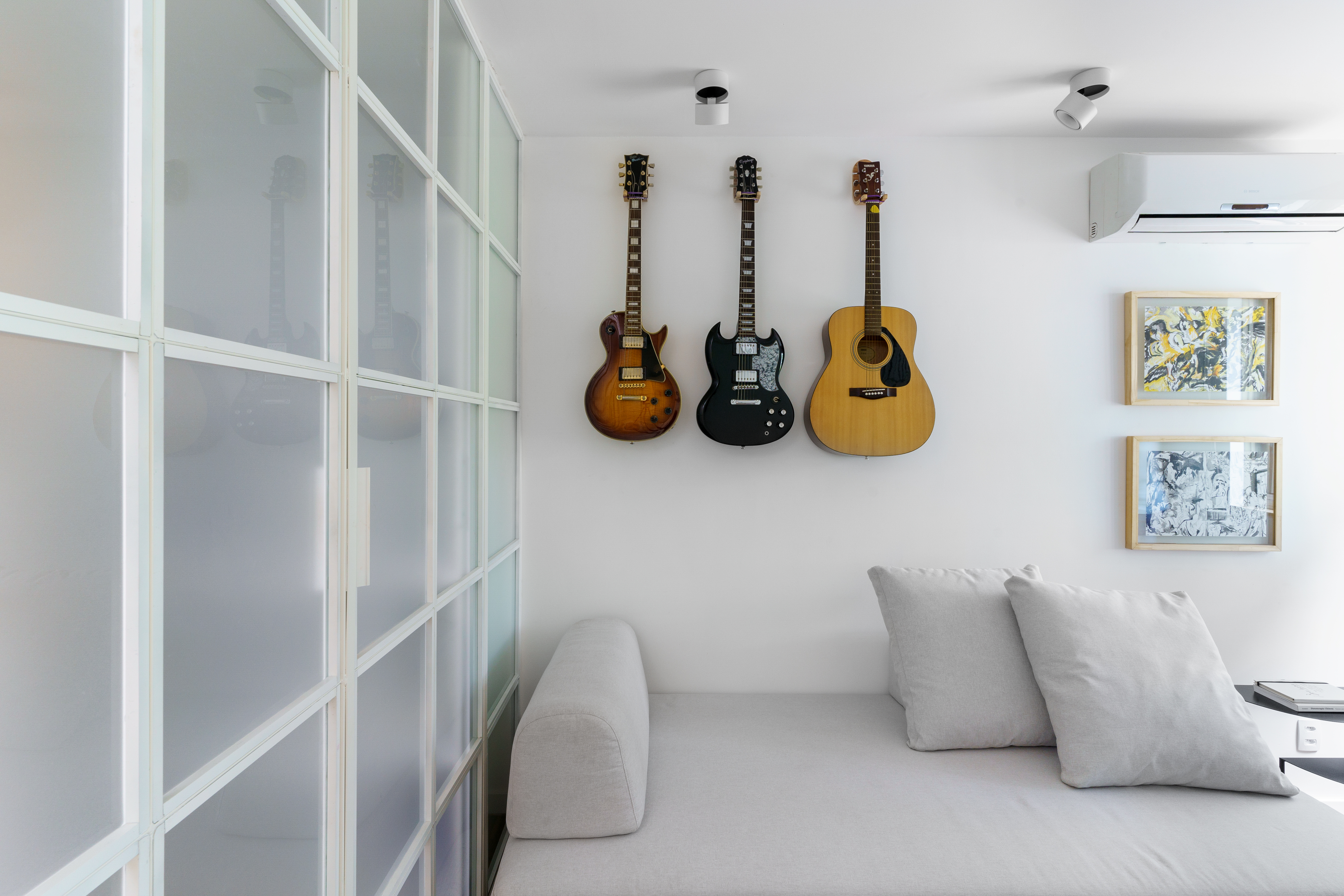 Projeto de Traama Arquitetura. Na foto, Quarto com sofá cama cinza claro, painel de vidro como divisória e instrumentos nas paredes.