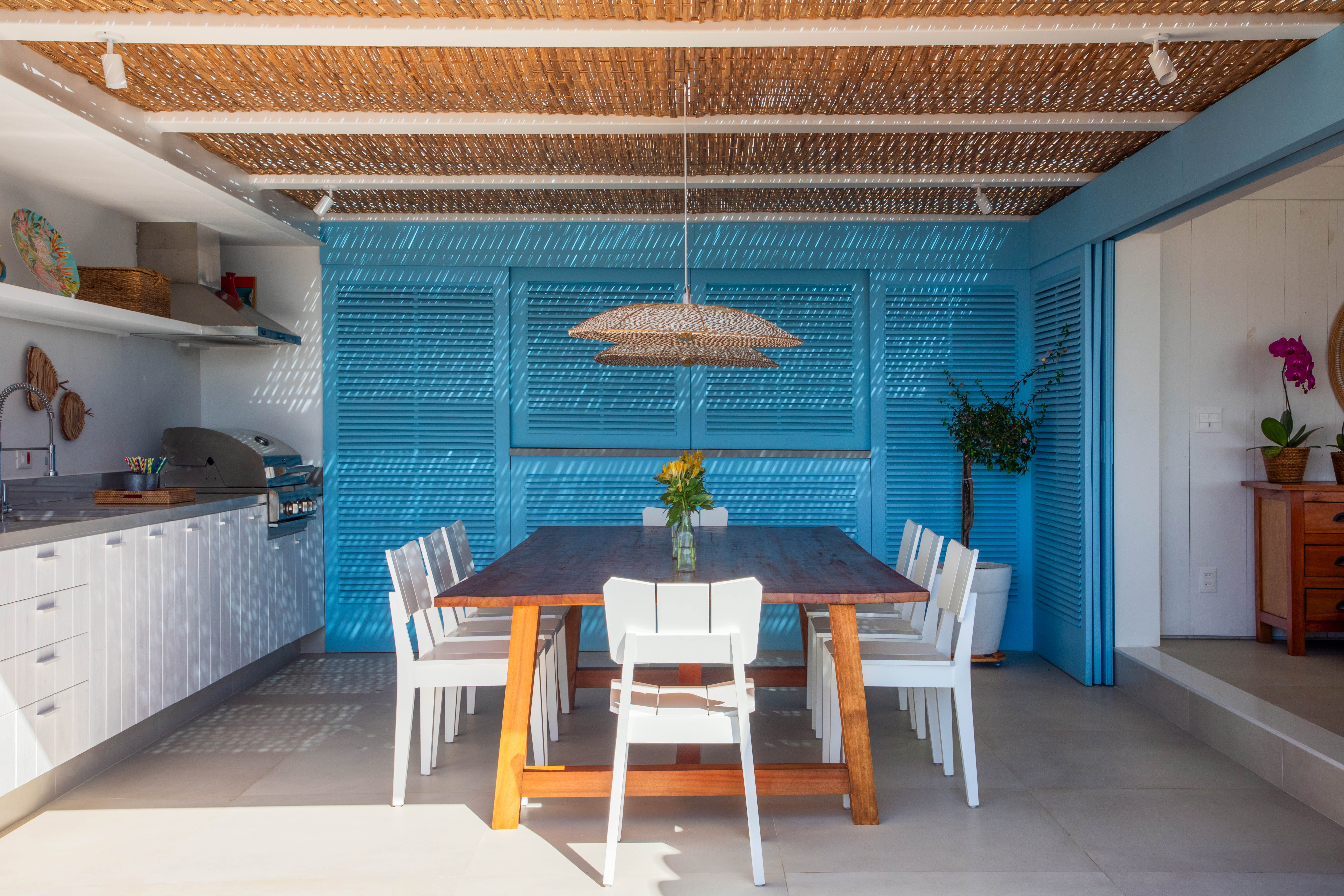 Projeto de Brise Arquitetura. Na foto, área gourmet com armários azuis, cobertura de pergolado de palhinha e mesa de madeira com cadeiras brancas.
