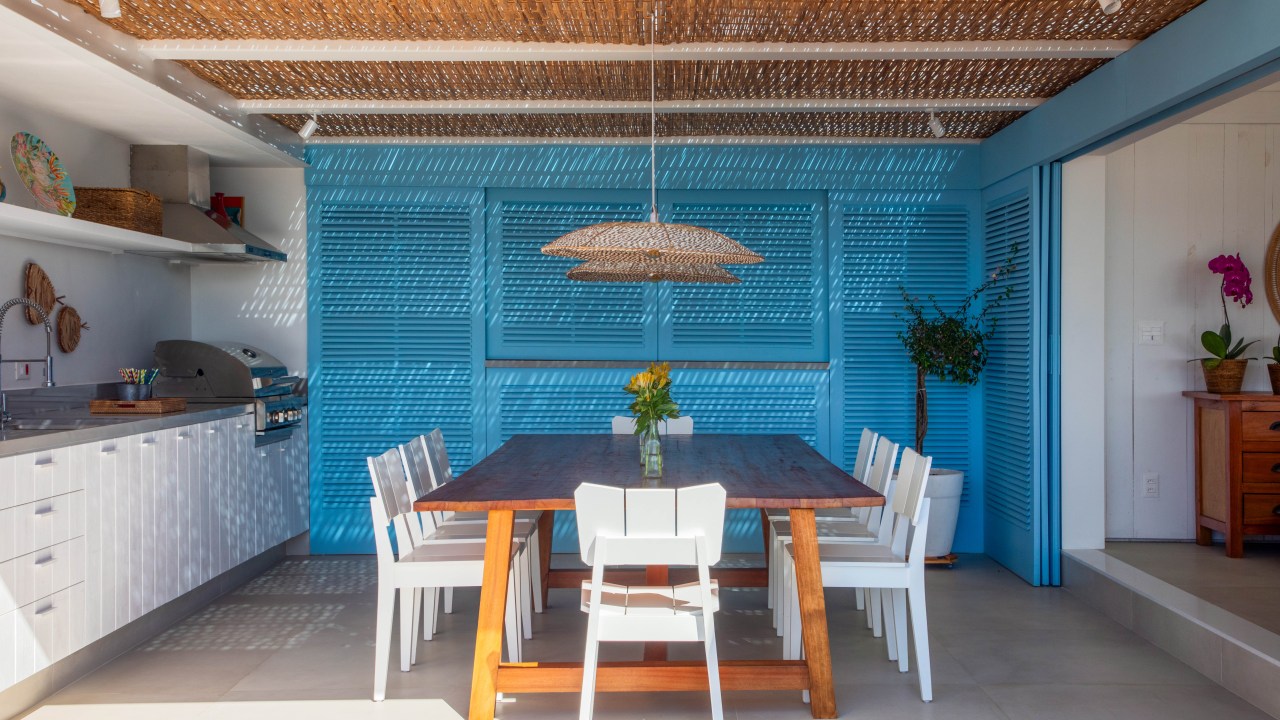 Projeto de Brise Arquitetura. Na foto, área gourmet com armários azuis, cobertura de pergolado de palhinha e mesa de madeira com cadeiras brancas.