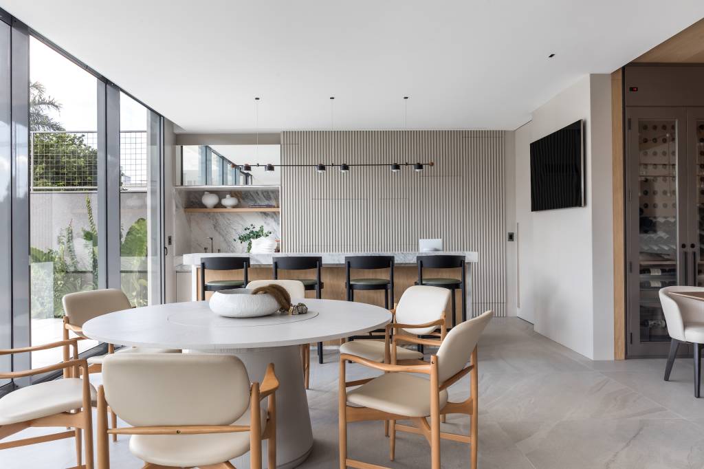 cores-neutras-design-clean-casa-450m-atemporal-larissa-loh-07-area-gourmet Projeto de Larissa Lóh. Na foto, área gourmet com mesa redonda de tampo branco, ilha com bancada e banquetas pretas.