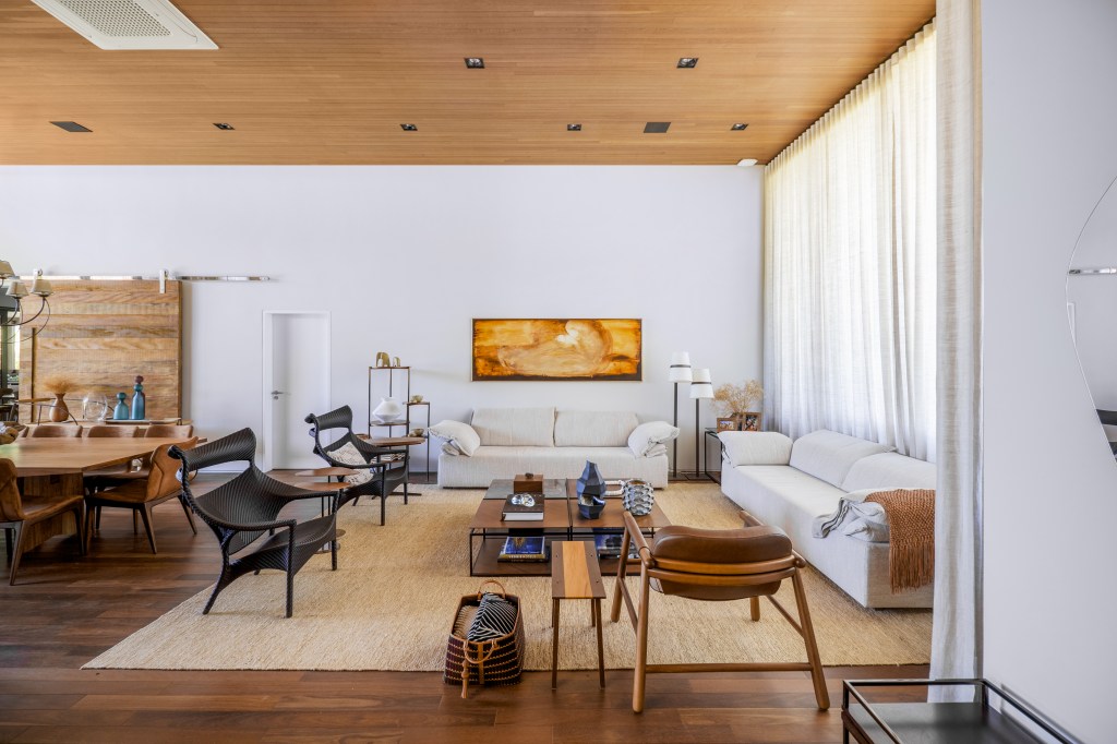 Projeto de TM Arquitetura. Na foto, sala de estar com decoração rústica com piso e teto de madeira, sofás brancos e cortina branca.