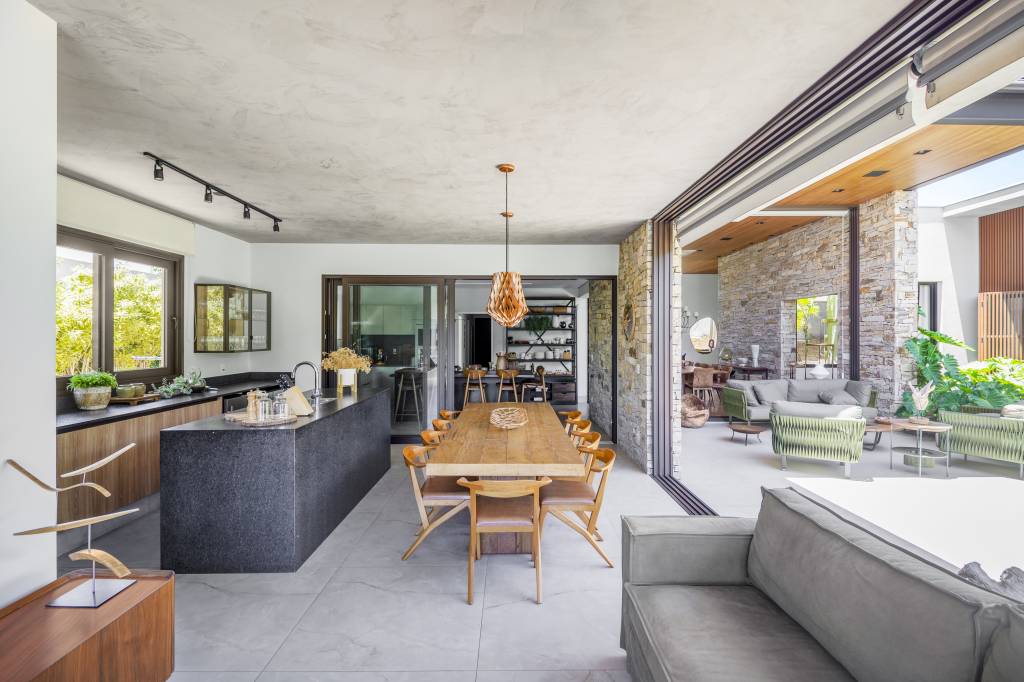 Projeto de TM Arquitetura. Na foto, área gourmet com piso de porcelanato, ilha revestida de granito preto, mesa rústica de madeira.