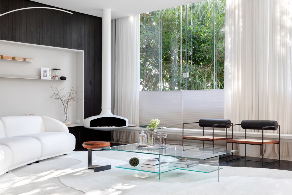 Madeira ebanizada é protagonista desta sala com décor branco e preto. Projeto de Barbara Ramos para a CASACOR Santa Catarina 2023. Na foto, sala de estar com lareira, sofá curvo e parede preta.