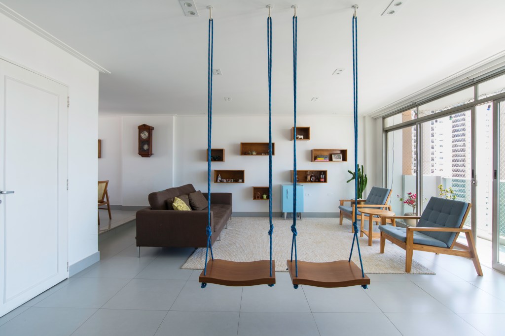Projeto de Raízes Arquitetos. Na foto, sala de estar com piso de porcelanato, sofá marrom, tapete cinza e balanços.