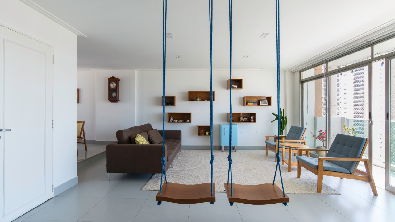 Projeto de Raízes Arquitetos. Na foto, sala de estar com piso de porcelanato, sofá marrom, tapete cinza e balanços.