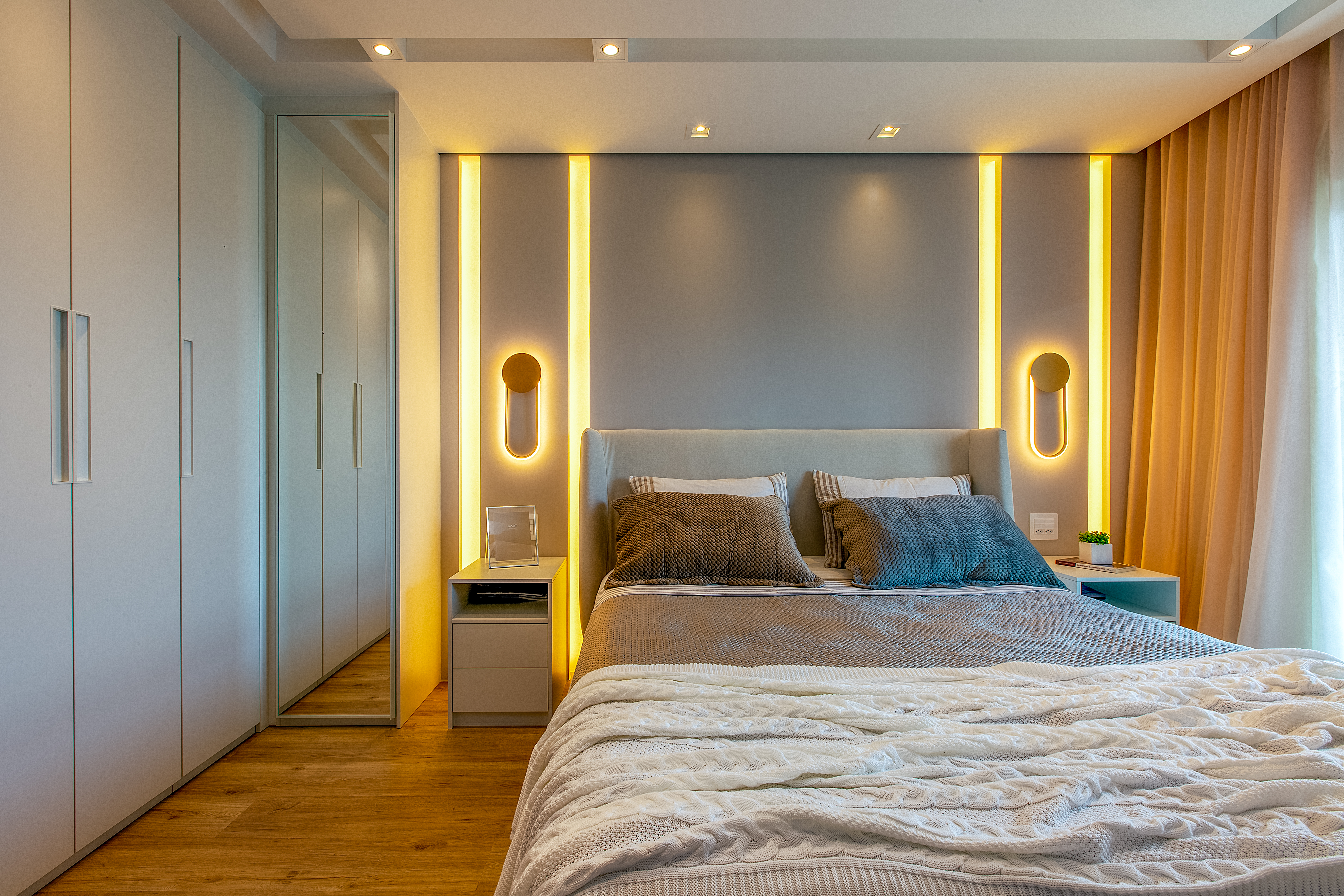 Projeto de Luciana Patriarcha. Na foto, quarto com cabeceira curva acolchoada e iluminação de LED. Arandelas ao lado da cama.