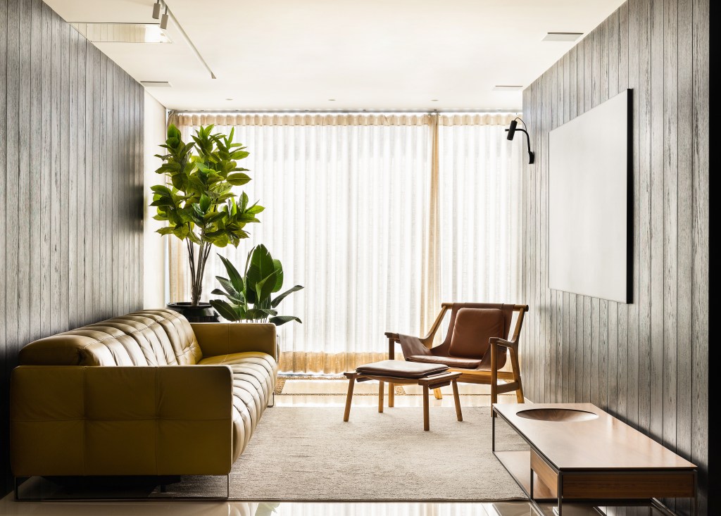 Painéis de carvalho ebanizados revestem as paredes deste apê de 320 m². Projeto de Lucas Lage. Na foto, sala de TV com parede de madeira, sofá e plantas.