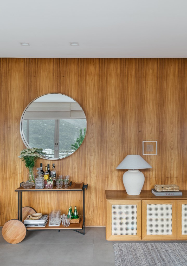 Projeto de A+G Arquitetura. Na foto, sala de jantar com parede revestida de madeira, carrinho de chá e espelho redondo.