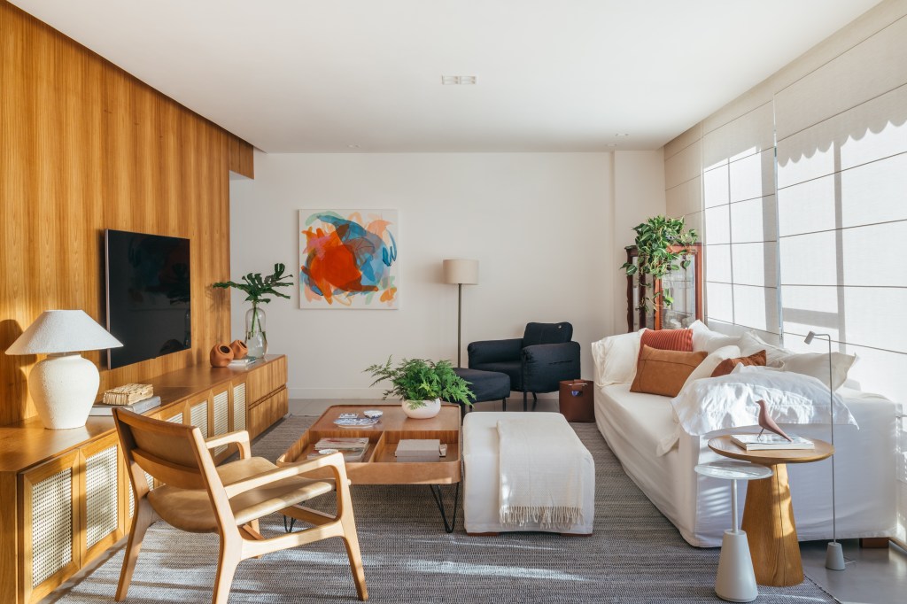 Projeto de A+G Arquitetura. Na foto, sala de estar com painel de madeira para tv, sofá branco com pufe, mesa de centro de madeira e poltrona azul.