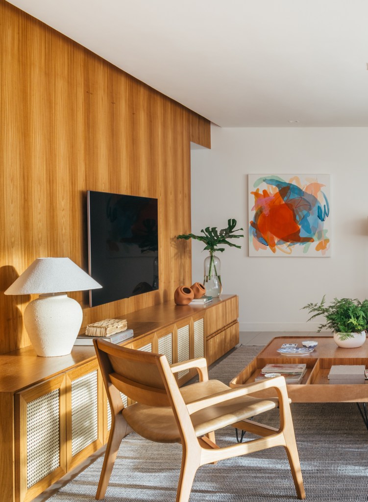 Projeto de A+G Arquitetura. Na foto, sala de estar com painel de madeira para tv, cadeira de madeira, mesa de centro e quadro colorido.