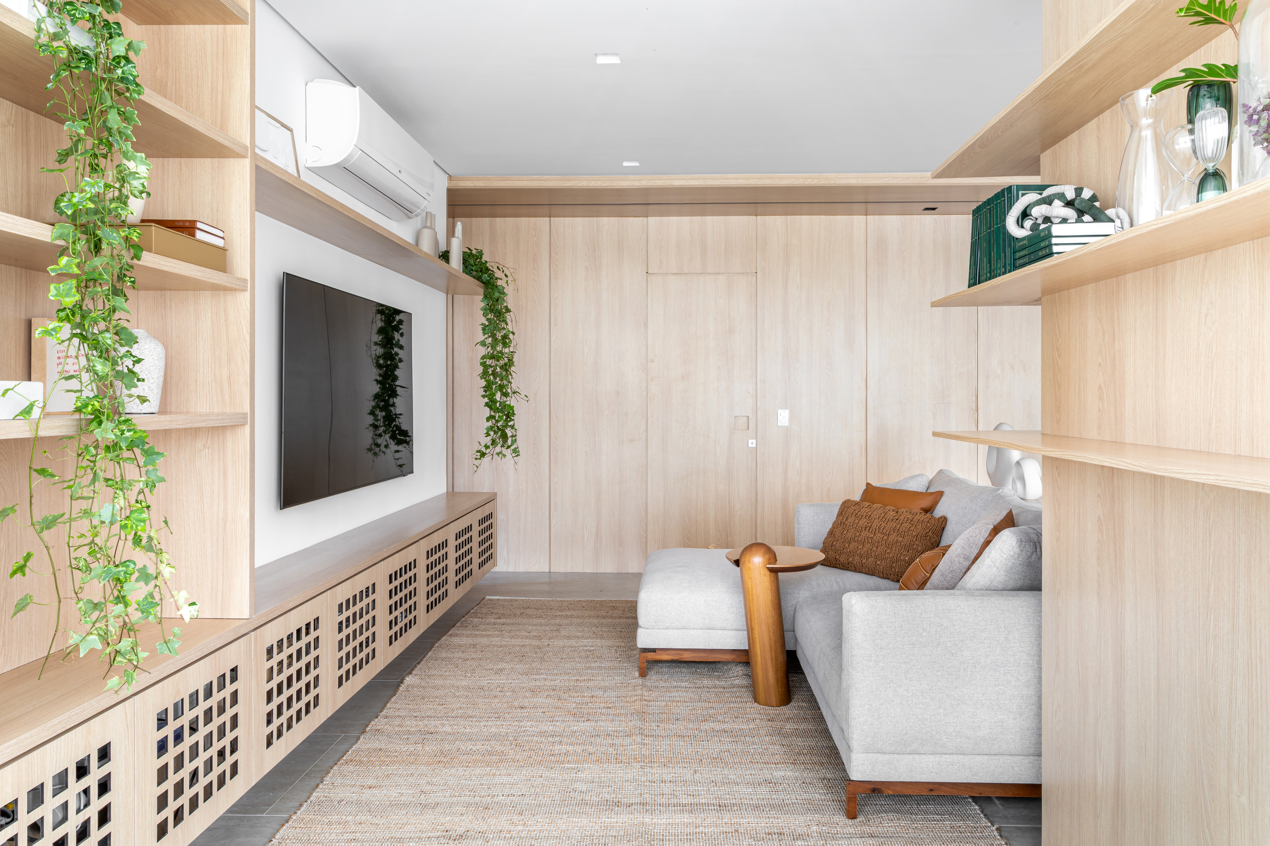 Projeto de NOP Arquitetura. Na foto, sala de estar com parede revestida de madeira clara, rack vazado e sofá cinza claro.