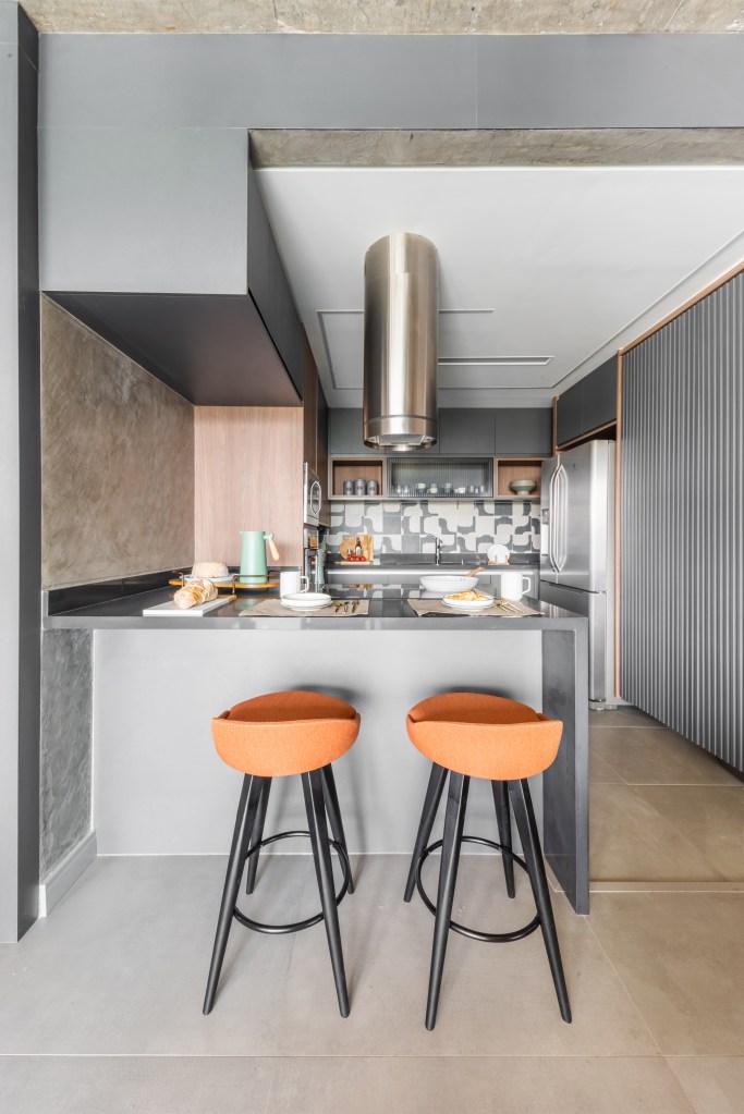 Projeto de BMA Studio. Na foto, cozinha pequena integrada com marcenaria cinza e bancada cinza com banquetas laranjas.