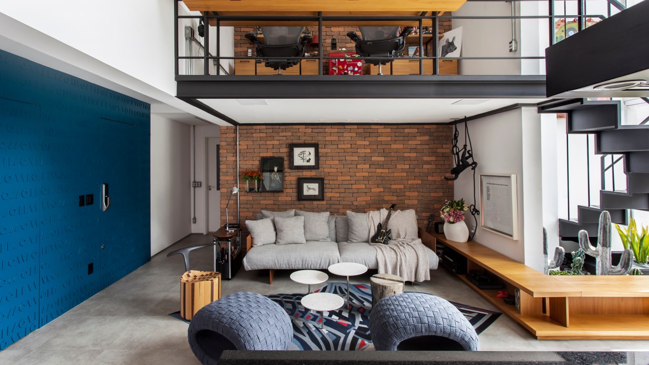 Projeto de Korman Arquitetos. Na foto, loft industrial com mezanino com home office e sala com sofá cinza e parede de tijolinhos.