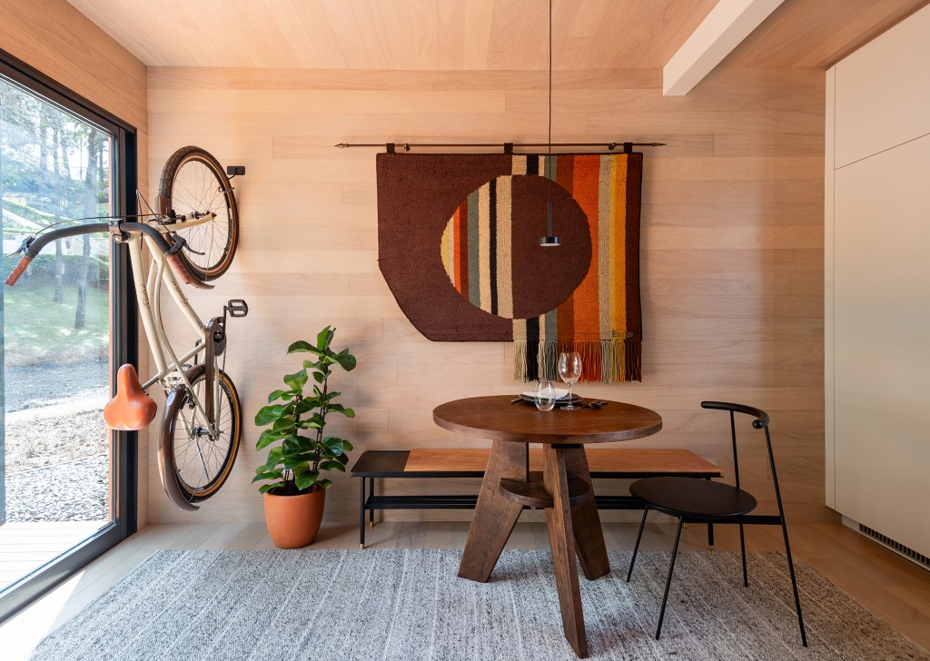 Tiny house sustentável de madeira fica pronta em 1 mês. Na foto, sala de jantar com tapecaria na parede e bicicleta.