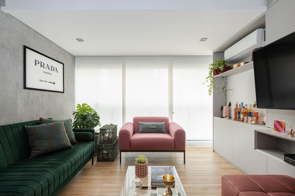 Projeto de Inovando Arquitetura. Na foto, sala de estar com parede de cimento queimado, sofá verde escuro, costela de adão e poltrona rosa.