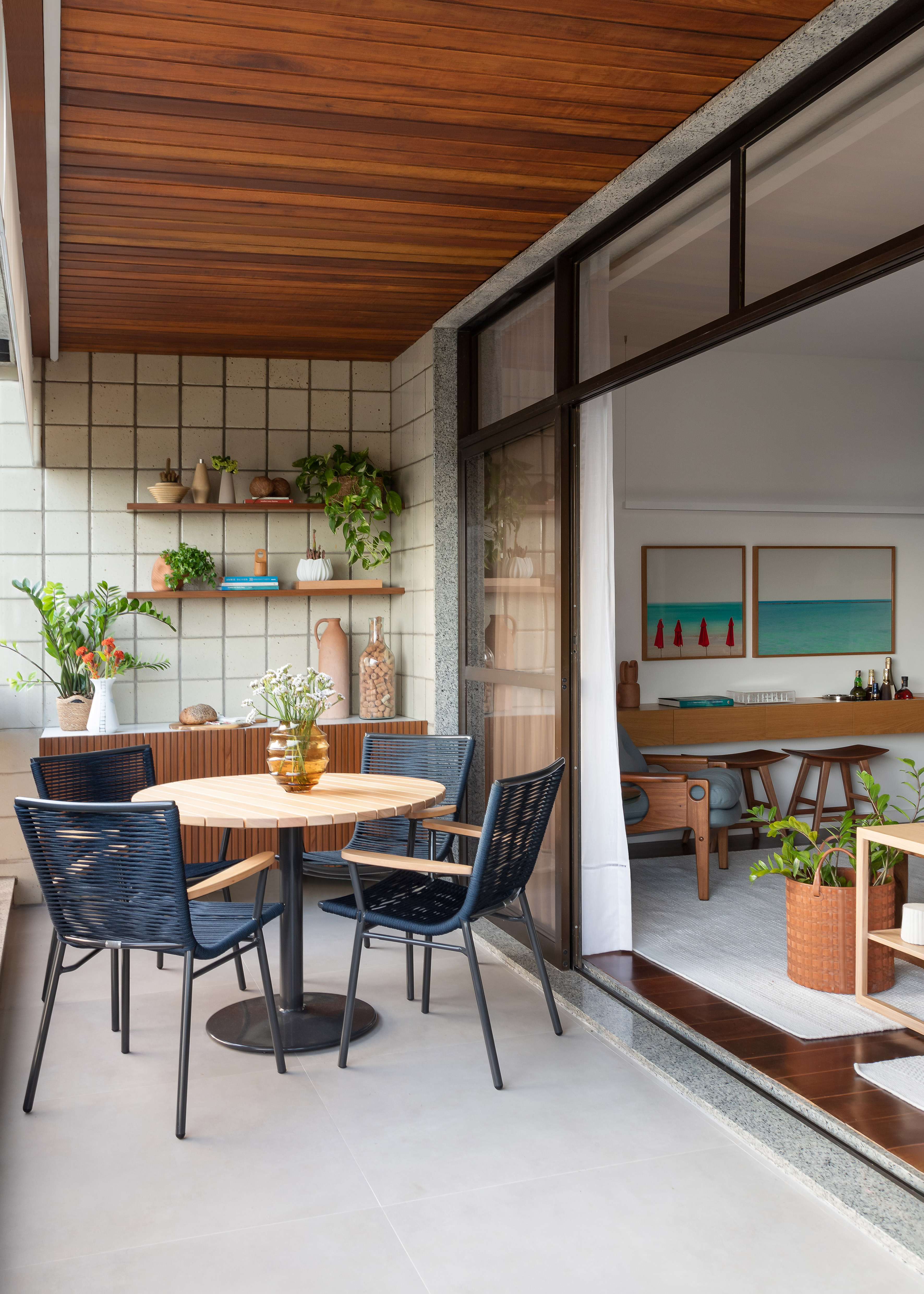 Projeto de MR Arquitetura. Na foto, varanda com mesa pequena redonda, cadeiras azuis e parede de azulejos com prateleiras com plantas.