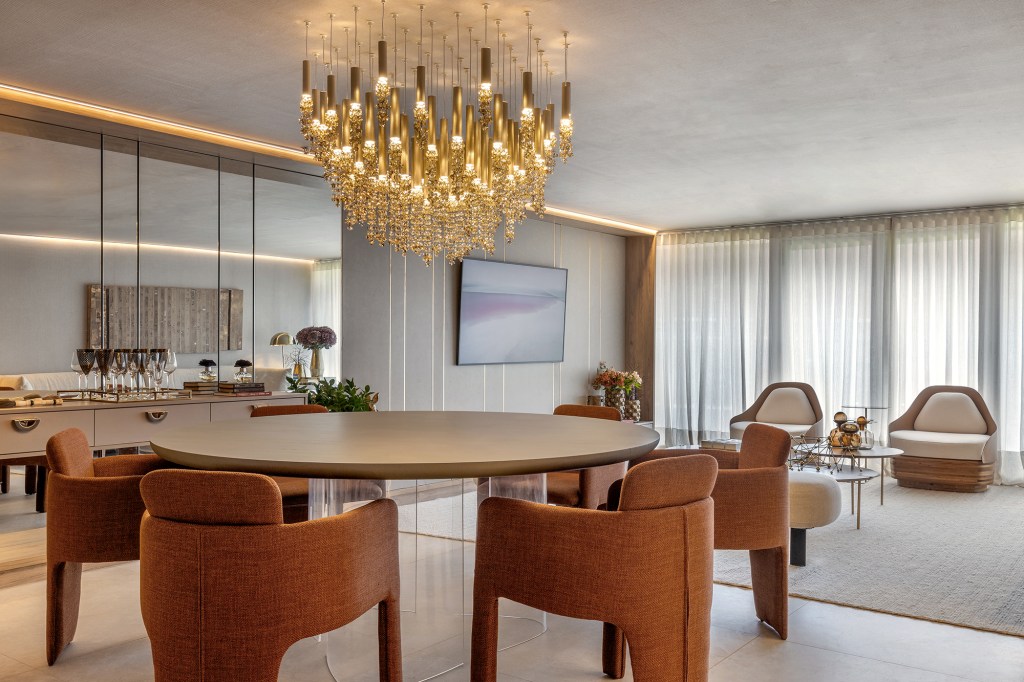 Apartamento de 189 m² tem décor afetivo e repleto de peças autorais. Projeto de Vivian Maia para a CASACOR Brasília 2023. Na foto, sala de estar e jantar com lustre de cristal, poltronas terrosas e espelho na parede.