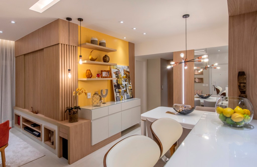 Apartamento de 85 m² tem décor baseada nas cores do chackras. Projeto de Campos & Olivetti Arquitetura. Na foto, sala estar com parede amarela e cozinha integrada,