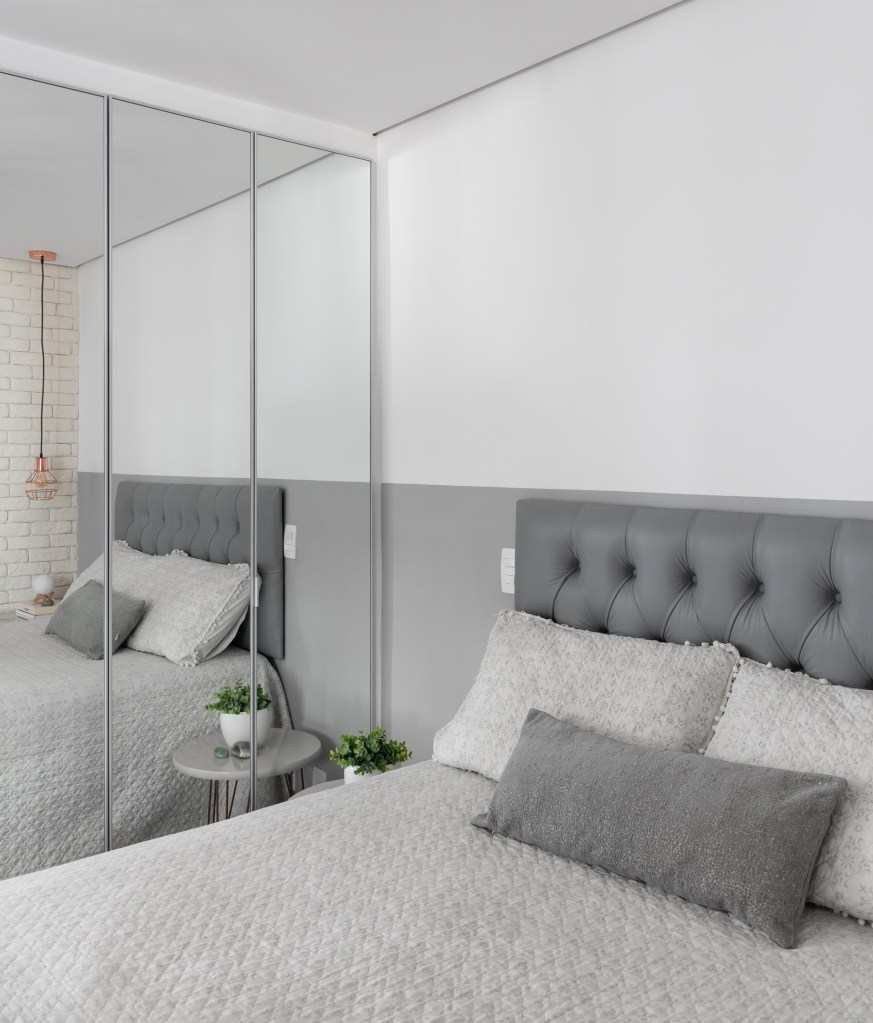 Apê de 31 m² em estilo escandinavo ganha amplitude com décor cinza e branco. Projeto de Estúdio Rena. Na foto, quarto de casal com cabeceira cinza em capitonê e armário espelhado.