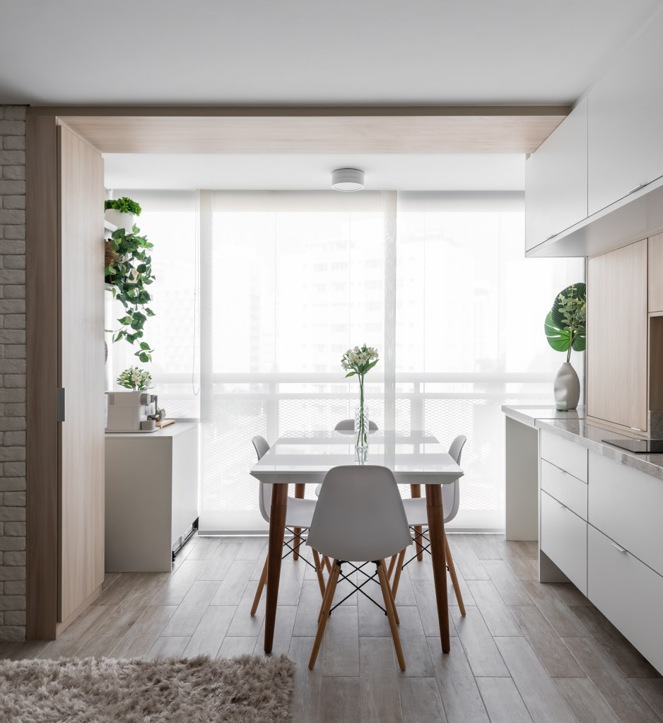 Apê de 31 m² em estilo escandinavo ganha amplitude com décor cinza e branco. Projeto de Estúdio Rena. Na foto, sala de jantar com mesa branca, cozinha integrada e parede de tijolinhos.