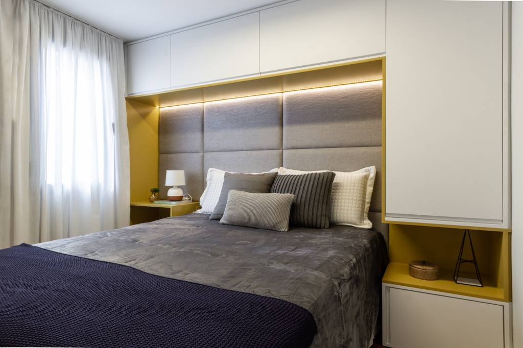 Apê de 53m² tem soluções pensadas para 3ª idade e quarto para netinha. Projeto de Mari Milan. Na foto, quarto de casal com marcenaria sobre a cama e cabeceira acolchoada cinza.