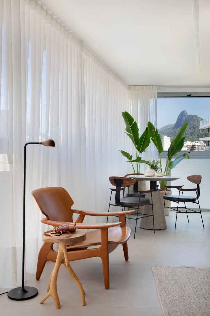 Apê neutro de 90m² aposta em porcelanato off white e cinza nos banheiros. Projeto de Rafael Ramos. Na foto, varanda com cadeira de madeira e cortina branca.