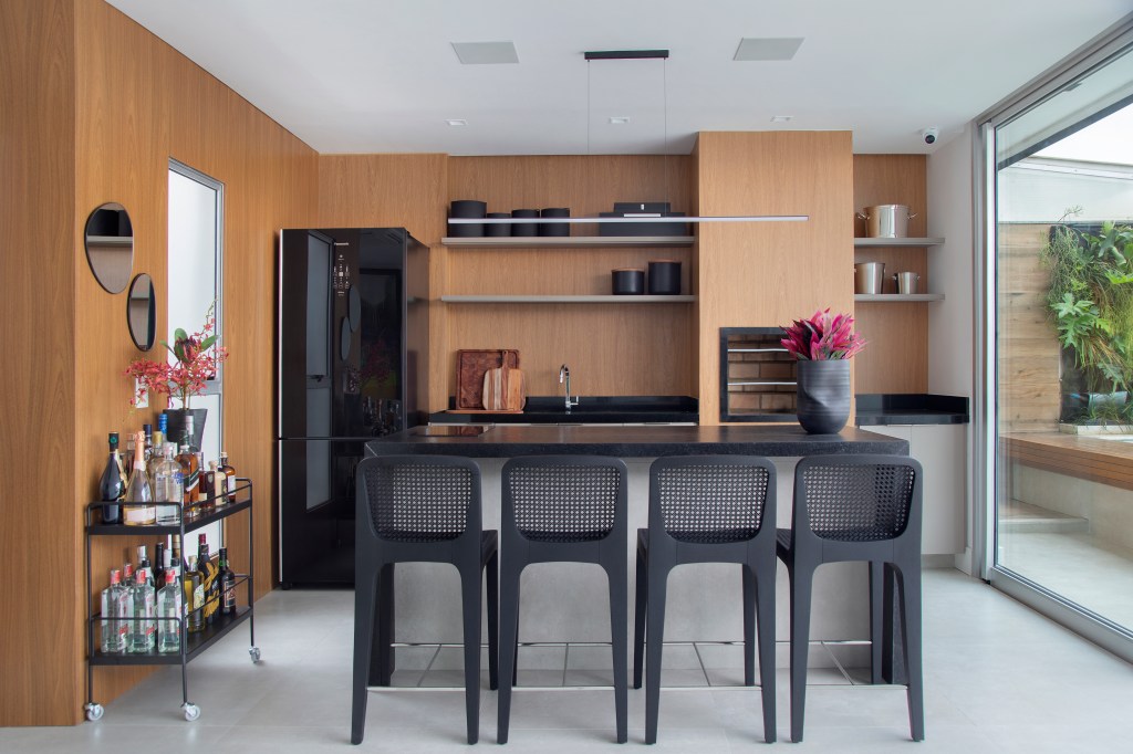Cobertura de 400m² para irmãos gêmeos tem closet espaçoso de 32m². Projeto de Tom Castro. Na foto, cozinha clean com paredes revestidas de madeira, bancada e banquetas pretas.