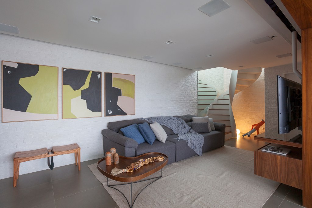 Dúplex de 150 m² ganha área externa com jacuzzi e escada escultural. Projeto de Felipe Carolo. Na foto, sala de estar com escada, sofá cinza e quadros.