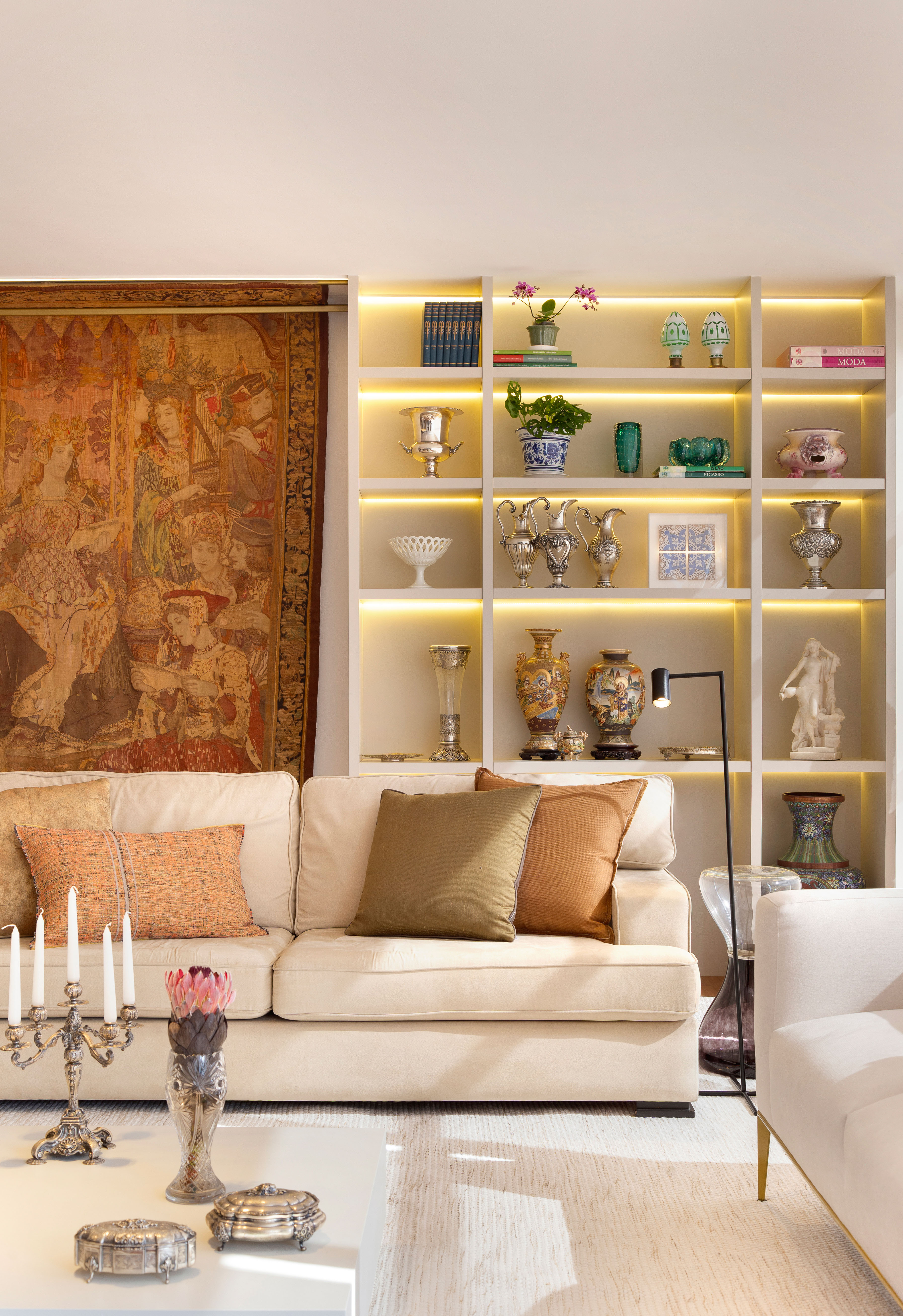 Projeto de Ricardo Melo e Rodrigo Passos. Na foto, sala de estar com sofá branco, tapete e estante de nichos iluminados.