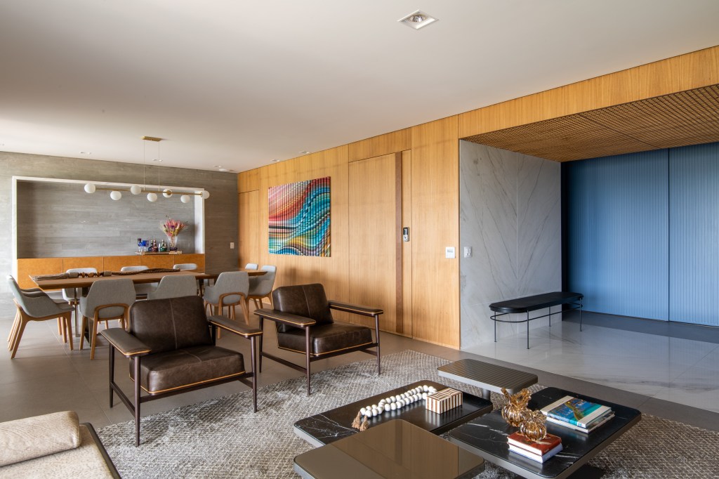 Forro iluminado de muxarabi cria sensação de luz natural em apê de 278 m². Projeto de Cris Furlan. Na foto, sala de estar com parede de madeira. Parece ripada azul. Jantar e quadros.