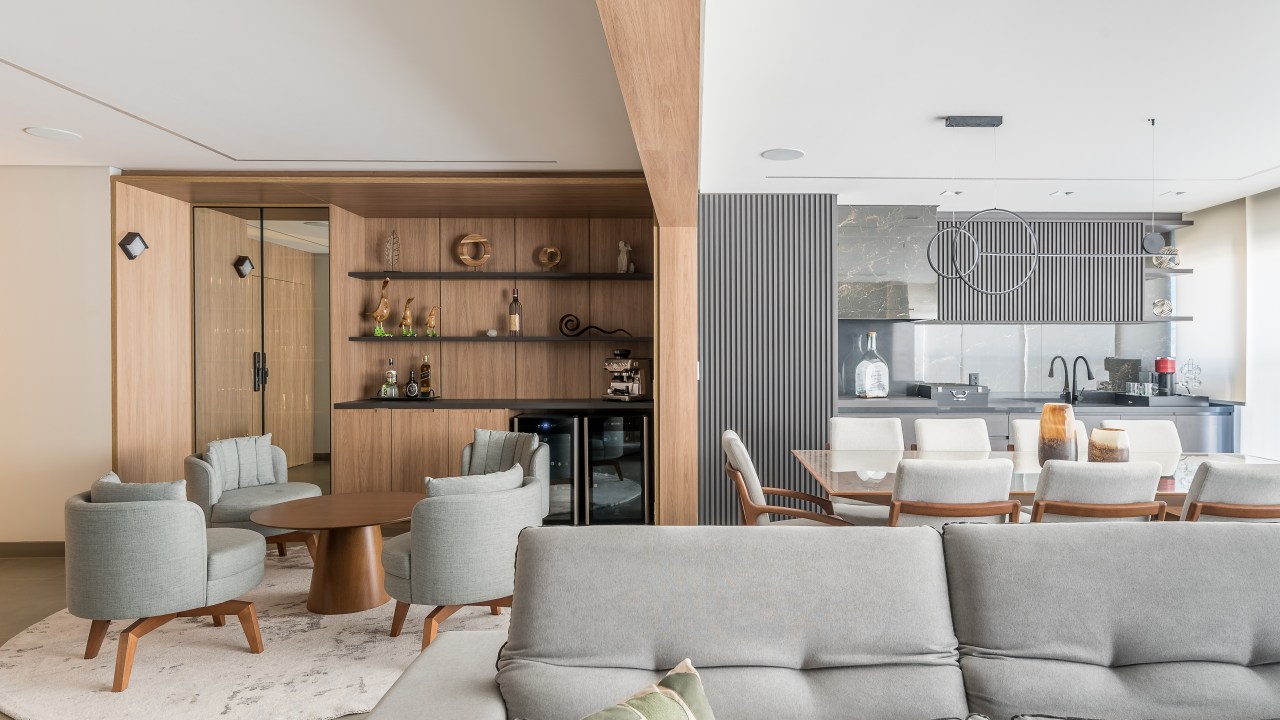 Marcenaria e sofá de 4m são destaques em apê aconchegante de 159m². Projeto de Elvira Oliveira Arquitetura. Na foto, sala de estar integrada com sofá cinza e adega.