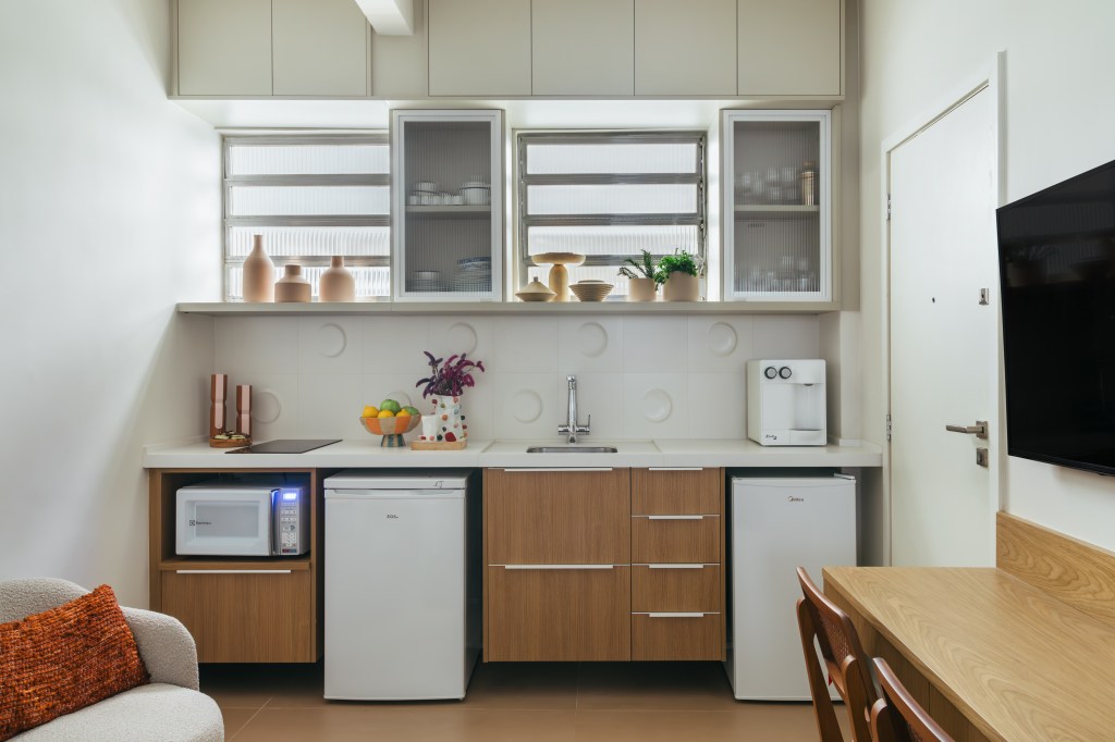 Projeto de Casa Cururu Arquitetura. Na foto, cozinha pequena com bancada branca, frigobar e armários brancos com portas de vidro.