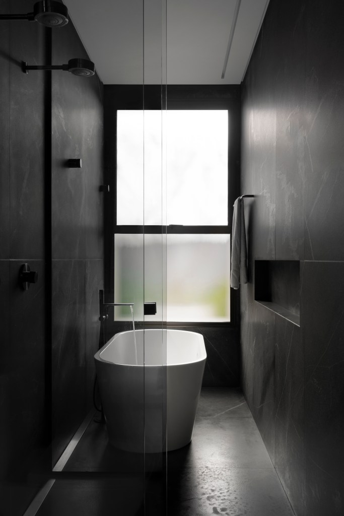 Minimalismo e décor contemporâneo marcam casa térrea de 273m². Projeto de Piacesi Arquitetos. Na foto, banheiro com revestimentos pretos e banheira solta branca.