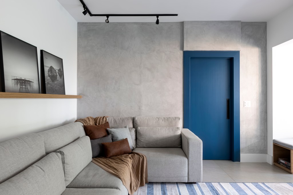 Com orçamento baixo, apê de 70m² ganha móveis planejados e churrasqueira. Projeto de Studio Monfré. Na foto, sala de estar com parede em cimento queimado, sofá cinza e porta azul.