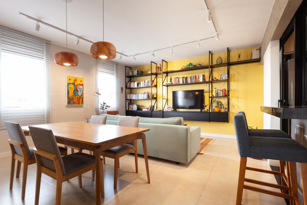 Cada parede sala é de uma cor neste apê de 80 m² com dois home offices. Projeto de Mutabile Arquitetura. Na foto, sala de estar e jantar com parede amarela, pendentes e sofá verde.