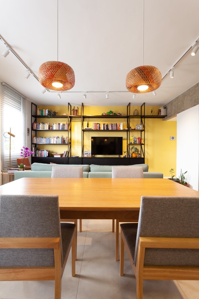 Cada parede sala é de uma cor neste apê de 80 m² com dois home offices. Projeto de Mutabile Arquitetura. Na foto, sala de estar e jantar com parede amarela, pendentes e sofá verde.