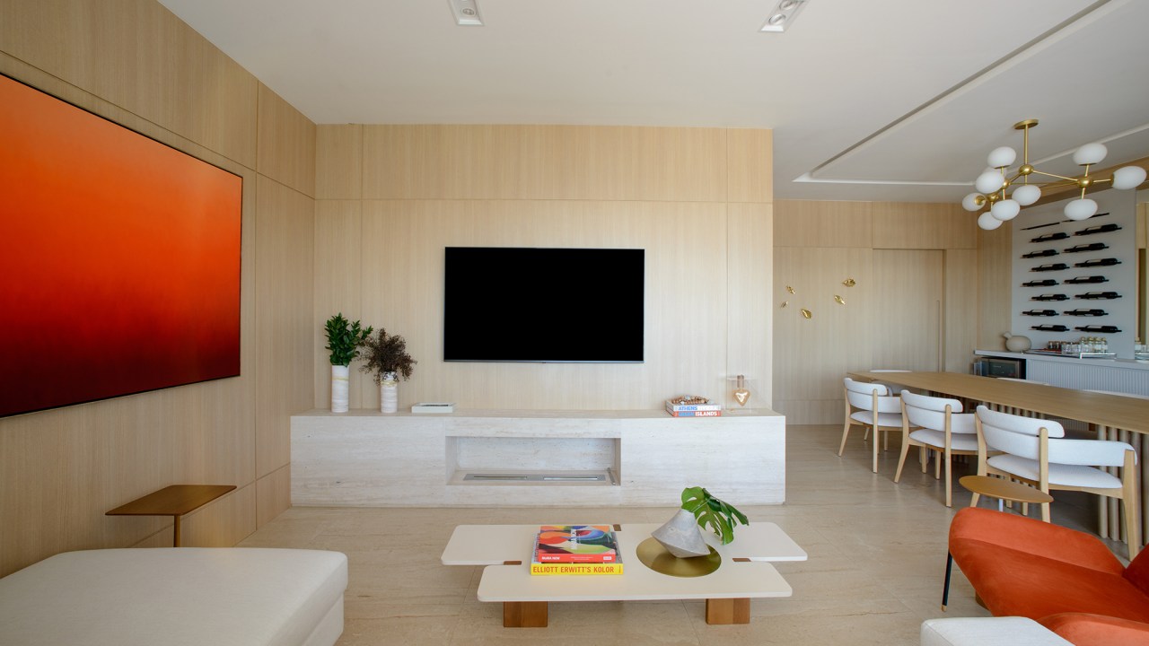 Paredes de madeira e tons de laranja compõem o décor deste apê de 138 m². Projeto de Letícia Nannetti. Na foto, sala de estar e jantar com parede de madeira, quadro e adega na parede.